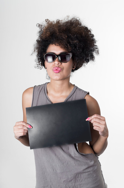 Hispanic brunette rebel model with afro like hair wearing grey sleeveless shirt and sunglasses making kiss lips holding up blank board as posing for mugshot concept - Foto, afbeelding