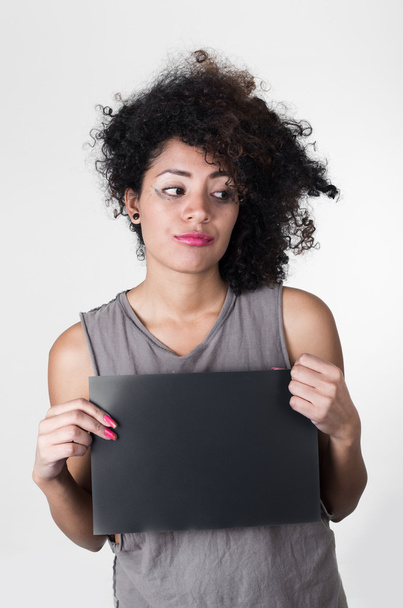Hispanic brunette model met afro zoals haar het dragen van grijze mouwloos shirt bedrijf leeg bord als poseren voor mugshot concept - Foto, afbeelding