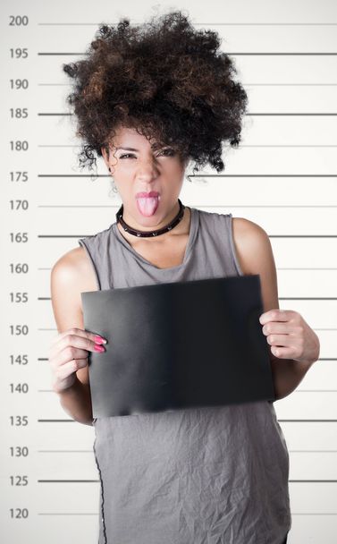 Hispanic brunette rebel model with afro like hair wearing grey sleeveless shirt holding up blank board as posing for mugshot concept and showing tongue to camera - Фото, зображення