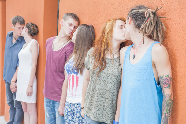 Keskikokoinen ihmisryhmä suutelee ja seisoo lähellä punaista seinää tausta
 - Valokuva, kuva