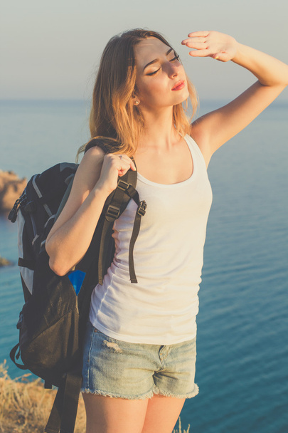 Happy backpacker girl is standing on rock over ocean view - Фото, изображение