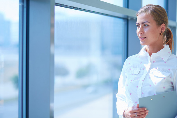 Businesswoman standing against office window holding documents in hand - Photo, Image