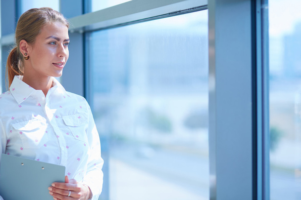 Businesswoman standing against office window holding documents in hand - Photo, image