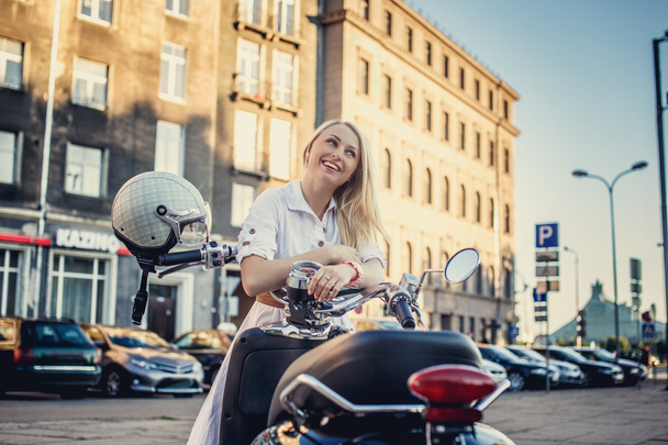 Усміхнена блондинка в білій сукні позує біля мотоциклетного скутера
. - Фото, зображення