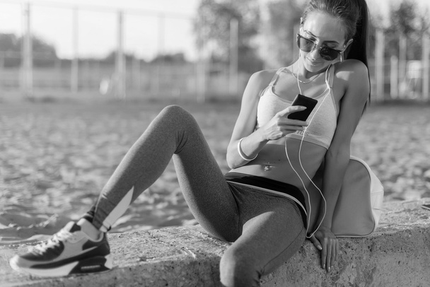 Beautiful fitness athlete woman wearing sunglasses resting listening music after work out exercising on summer evening in beach at sunset with smartphone outdoor portrait. - Photo, Image