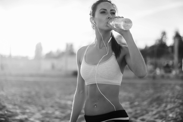 Hermosa atleta de fitness mujer descansando agua potable después de hacer ejercicio en la playa noche de verano en sol soleado retrato al aire libre
 - Foto, imagen