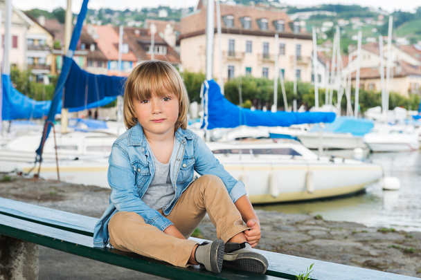 Portrét móda roztomilý malý blonďák sedí na lavičce u jezera, na sobě džínové košili a béžové kalhoty - Fotografie, Obrázek