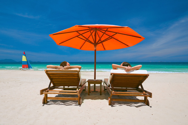 Couple on a tropical beach on deck chairs under a red umbrella - Photo, Image