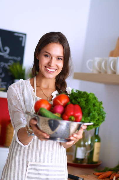 Mujer joven sonriente sosteniendo verduras de pie en la cocina - Foto, imagen
