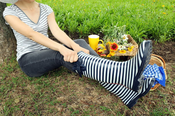 Young woman in rubber boots sitting on meadow outdoors - Foto, Imagen