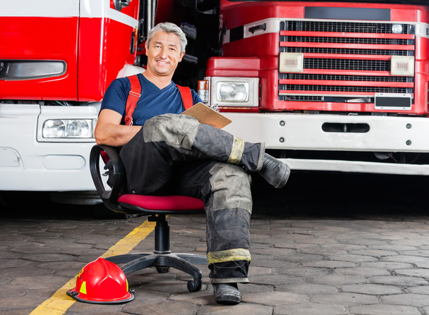 Happy Firefighter Sitting On Chair Against Trucks - Photo, Image