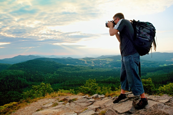 Photographer with big mirror camera on neck and backpack stay on peak of rock. Hilly landscape, fresh green color in valley. - Photo, Image