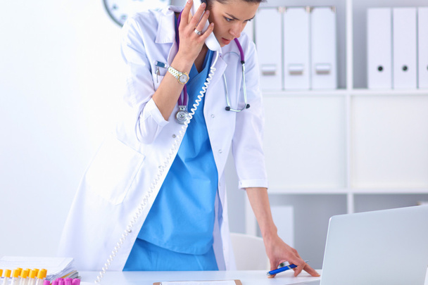 Young woman doctor in white coat at computer using phone - Фото, изображение