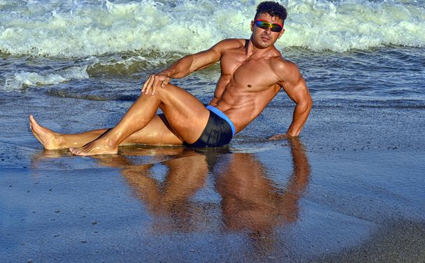 Strong bodybuilder with six pack.Fitness trainer with perfect abs, shoulders,biceps, triceps,chest, flexing his muscles on the beach with sea waves on the background, training in vacation - Φωτογραφία, εικόνα
