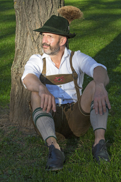 man in traditional bavarian clothes sitting by a tree - Foto, imagen