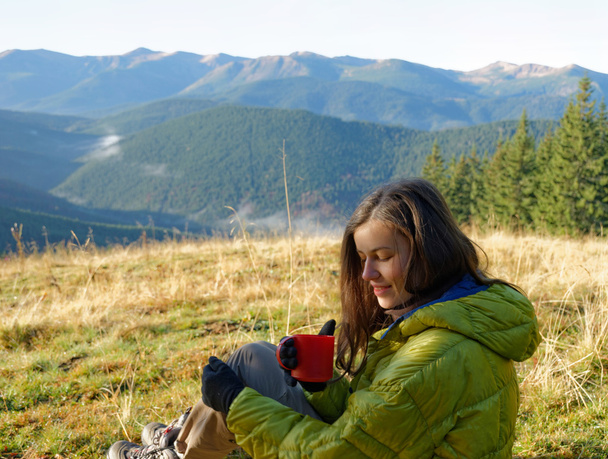 Portrait of girl in yellow down jacket sitting with red cup of hot beverage in the autumn mountains with mist in the valleys - Valokuva, kuva