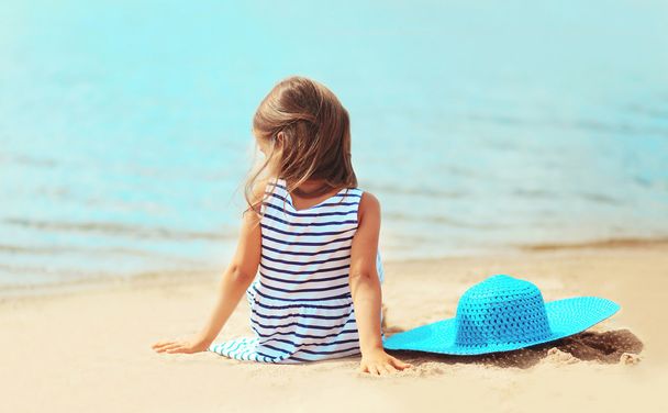 Little girl sitting on the sand beach with straw hat near sea in - Foto, Bild
