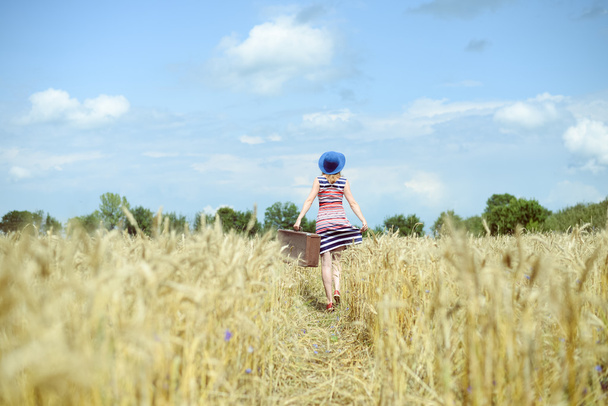 Woman wearing hat with suitcase walking away through wheat field - Fotografie, Obrázek