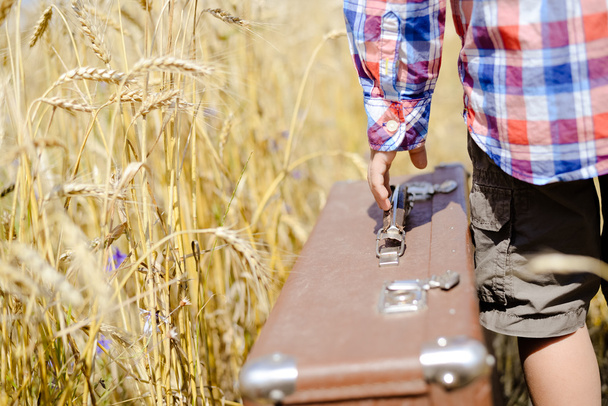 Boys hand with big old retro suitcase in wheat field over sunny outdoors background - Фото, зображення