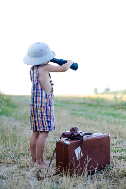 Kid wearing pith hat and plaid romper looking in spyglass near big suitcase. - Foto, afbeelding