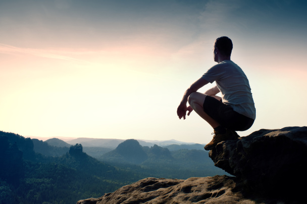 Hombre joven en pantalones deportivos negros y camisa gris está sentado en el borde del acantilado y mirando al valle brumoso abajo
 - Foto, imagen