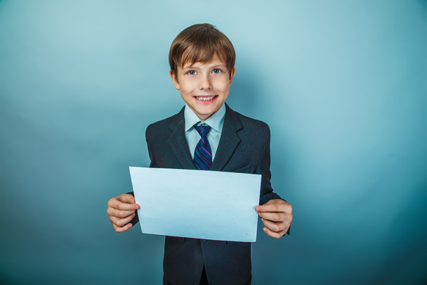 europäisches Aussehen Teenager Junge mit blonden Haaren in Anzug und Krawatte hält ein Blatt weißes Papier auf einem grauen Hintergrund, lächeln - Foto, Bild