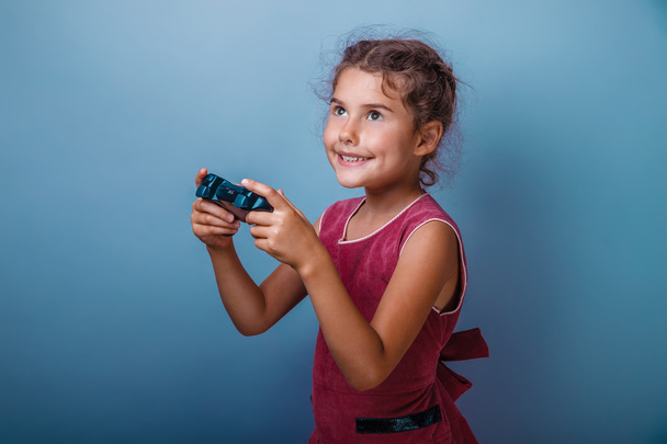 Girl seven years old, European-looking brunette in a pink dress holding a joystick and smiling on a gray background, happiness, joy, game - Photo, Image