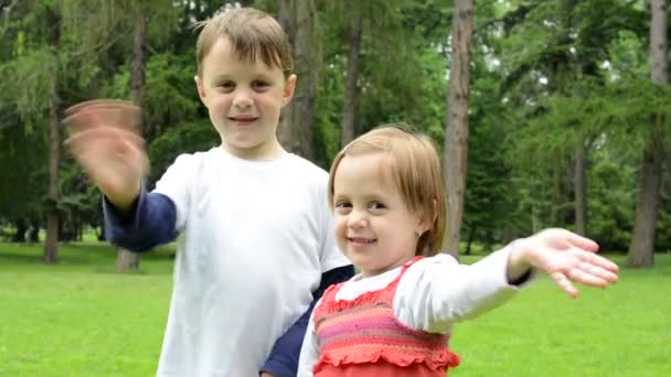 Kinder (kleiner Junge und kleines Mädchen (Geschwister) winken mit Händen und lächeln in die Kamera - Park - Filmmaterial, Video