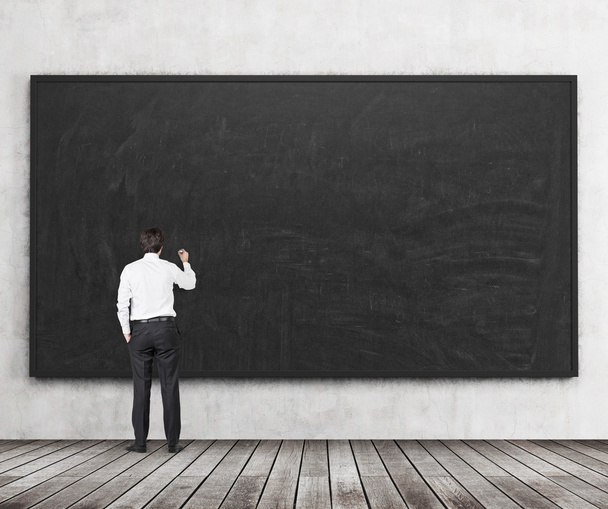 Rear view of the man who is going to write something on the black chalkboard. Wooden floor and concrete wall. A concept of the beginning of new academic year. A class room. - 写真・画像