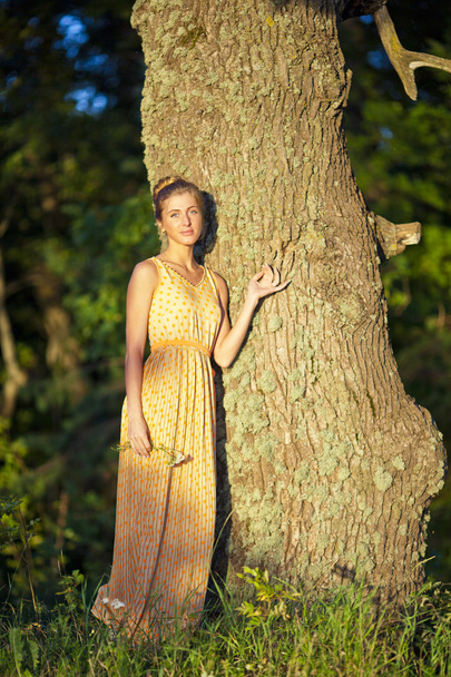 young girl in a yellow dress standing at the tree with a flower in the hands - Foto, Imagen
