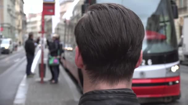 młody przystojny hipster człowiek patrzeć na opuszczenie tramwaju ze stacji w mieście - ulica miejska z ludźmi - Materiał filmowy, wideo