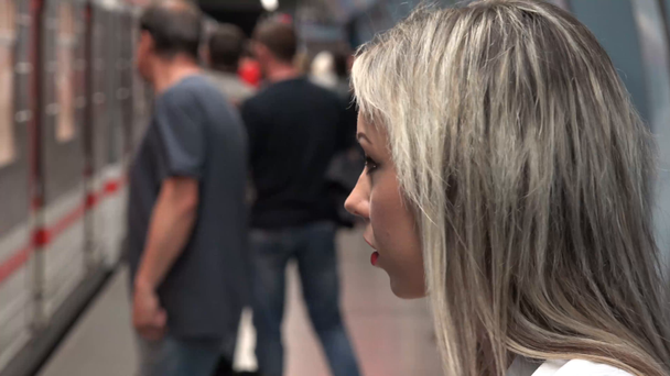 молодая привлекательная блондинка ждет метро и оглядывается вокруг - прибывает метро - люди входят и выходят из метро - женщина садится на поезд метро
 - Кадры, видео