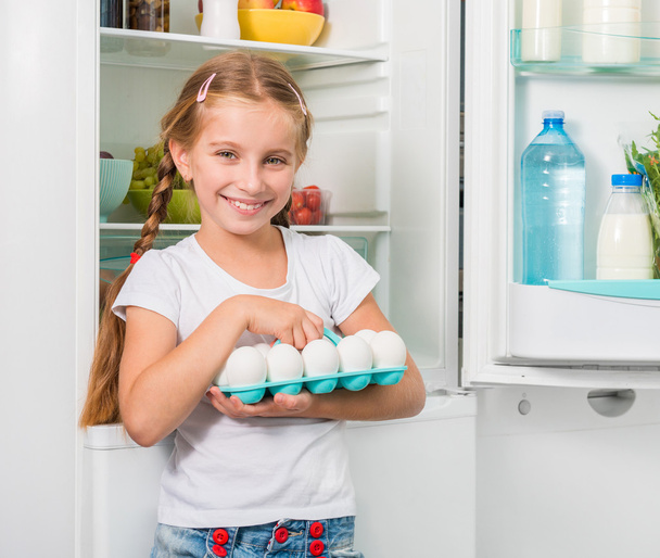 little girl holding eggs near fridge - Zdjęcie, obraz