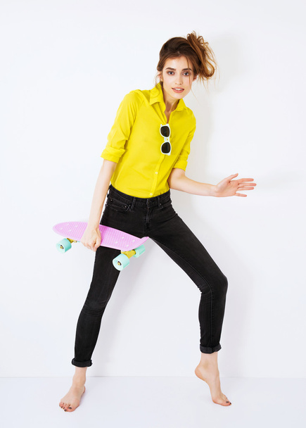 Mode fille hipster en chemise jaune dans les lunettes de soleil avec planche à roulettes flirter contre le mur blanc
. - Photo, image