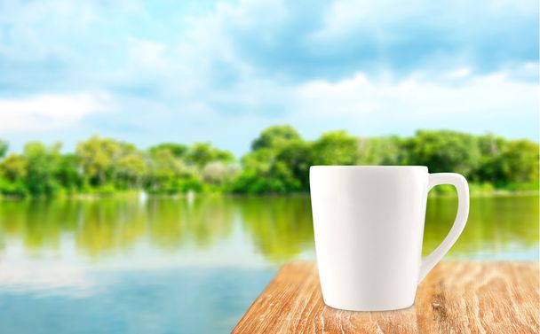 Біла чашка кави на дерев'яному столі на розмитому дереві та фоні лагуни
 - Фото, зображення