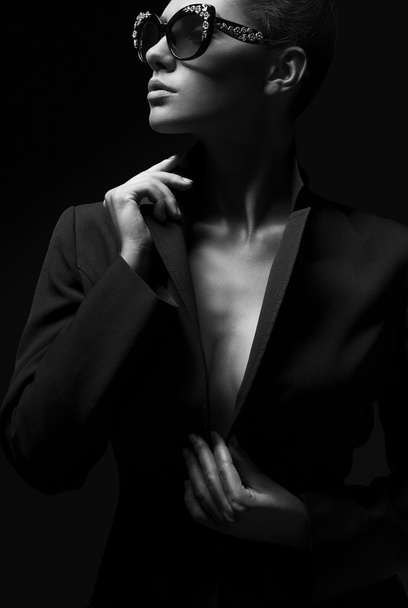 Porträt eines schönen Mädchens, das in schwarzer Jacke und modischer Sonnenbrille im Studio posiert. das Konzept der stilvollen und sexy Frauen. perfekte Haut und Körper. Schwarz-Weiß-Foto - Foto, Bild