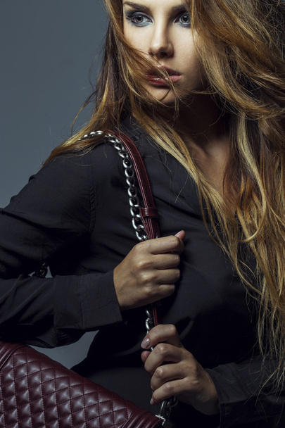 moda chica rubia sosteniendo bolso de mano de Borgoña con el pelo largo, maquillaje sexy y camisa negra posando sobre fondo gris
. - Foto, imagen