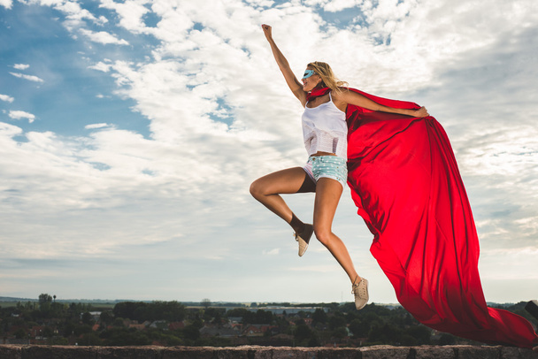 Mujer rubia en vestido rojo y manto rojo saltando al aire libre como un superhéroe contra el cielo azul
 - Foto, Imagen