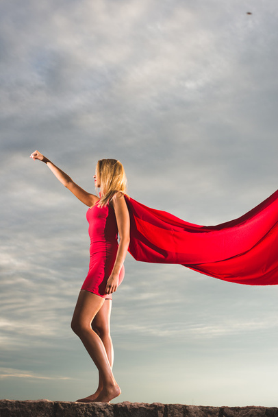 Szőke nő piros ruhában és piros köpeny jelentő szabadtéri, mint egy szuperhős kék ég ellen - Fotó, kép