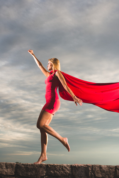 Blonde woman in red dress and red mantle posing outdoor as a superhero against blue sky - Photo, Image