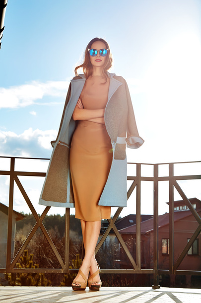 Beautiful fashionable young business woman wearing in ?rimson autumn coat and sunglasses with hairdo and makeup walking on a street with fall near big house and trees without yellow leaves background - Zdjęcie, obraz