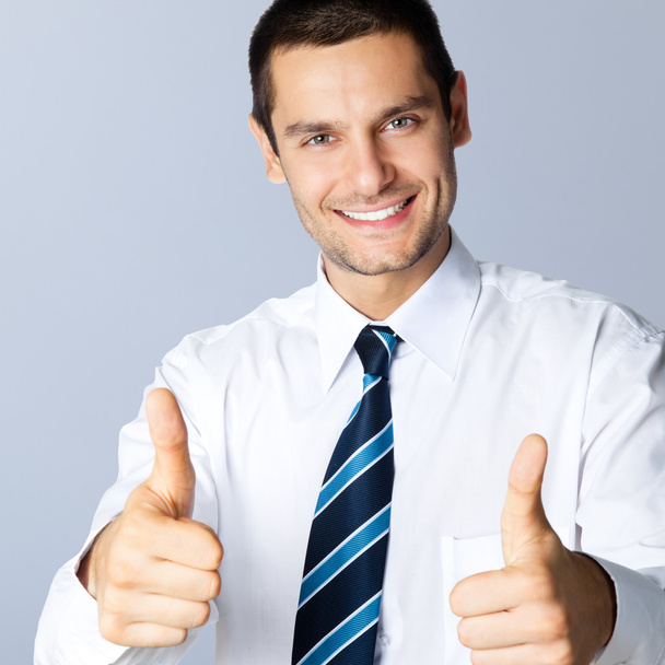Smiling businessman with thumbs up gesture, on grey - 写真・画像
