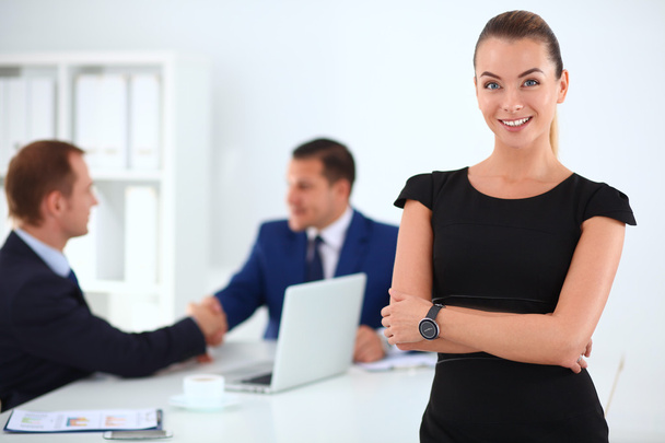 Portrait of a beautiful businesswoman standing in an office with colleagues shaking hands in the background - Foto, Imagem