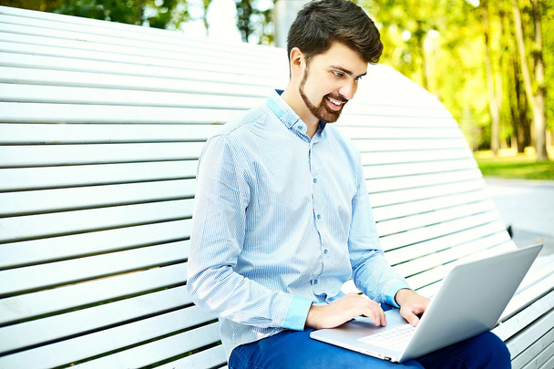Young handsome smiling businessman model sitting on the park bench using laptop in casual hipster cloth - Fotó, kép