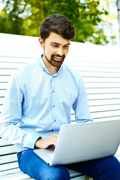 Young handsome smiling businessman model sitting on the park bench using laptop in casual hipster cloth - Zdjęcie, obraz