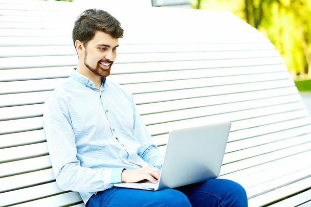 Young handsome smiling businessman model sitting on the park bench using laptop in casual hipster cloth - Фото, изображение