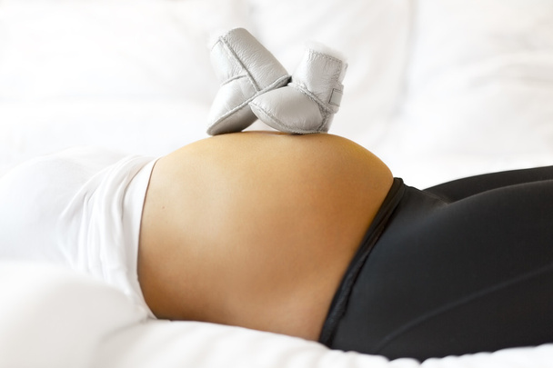 彼女の腹の赤ちゃんの靴が付いているベッドで妊娠中の女性 - 写真・画像