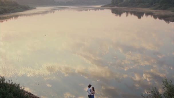 Mladý zamilovaný pár objímající se za úsvitu u jezera. Ranní snové rande v přírodě. Krásný odraz mraků v jezerních vodách. Nádherná vodní scenérie se dvěma lidmi stojícími na břehu řeky. Zpomalený pohyb - Záběry, video