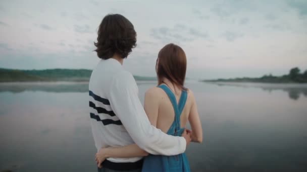 Young couple in love embracing together by the lake. Boyfriend and girlfriend holding each other in nature looking at lake water and waiting for sunrise. Slow motion - Footage, Video
