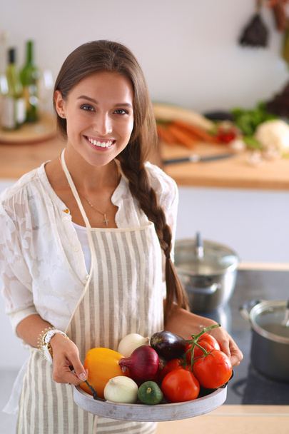 Smiling young woman holding vegetables standing in kitchen - Valokuva, kuva